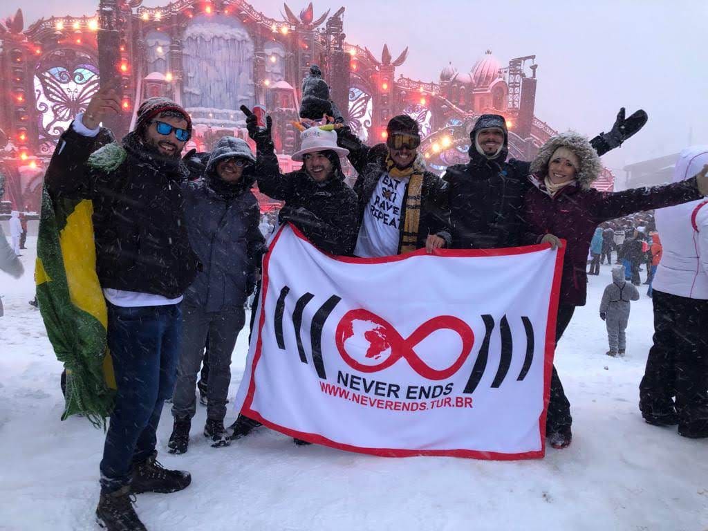 Grupo da Never Ends com a bandeira da agência aberta na neve, debaixo de nevasca, diante do palco principal da primeira Tomorrowland Winter em Alpe d'Huez