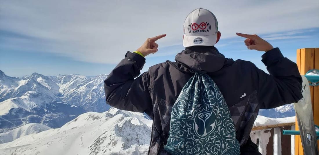 Viajante da Never Ends de boné da agência e mochila da Tomorrowland apontando pros picos nevados dos Alpes franceses, no alto da montanha de Alpe d'Huez
