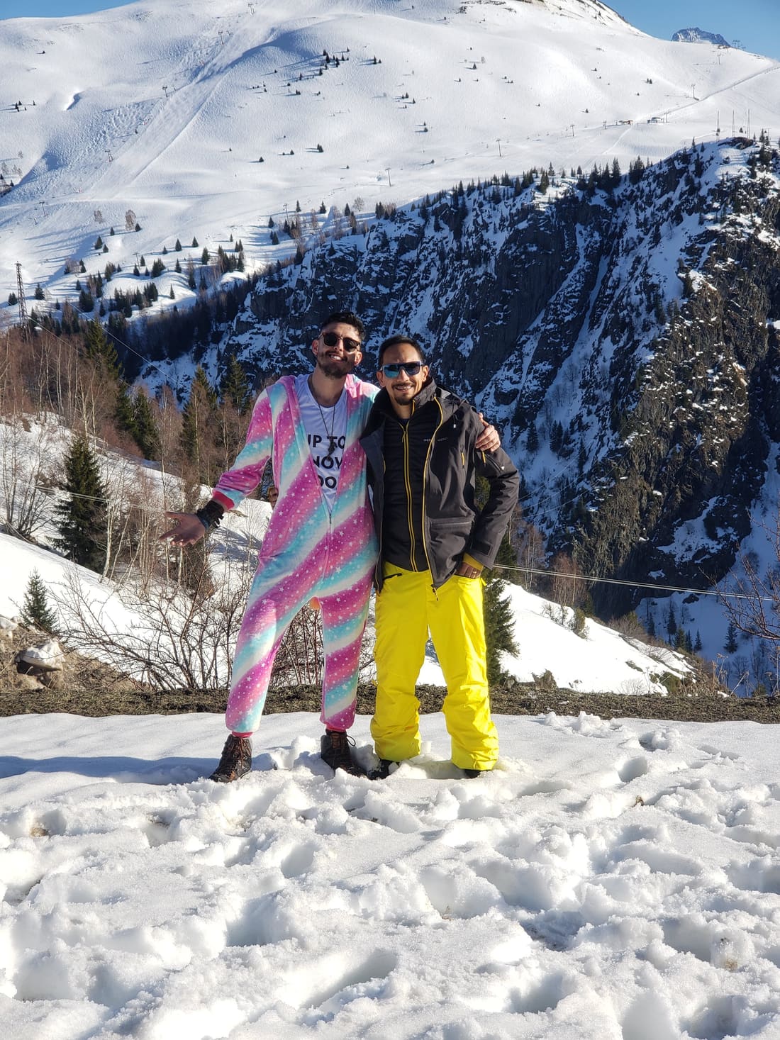 Dois viajantes da Never Ends posando na neve de Alpe d'Huez, com a pista de esqui e a montanha atrás