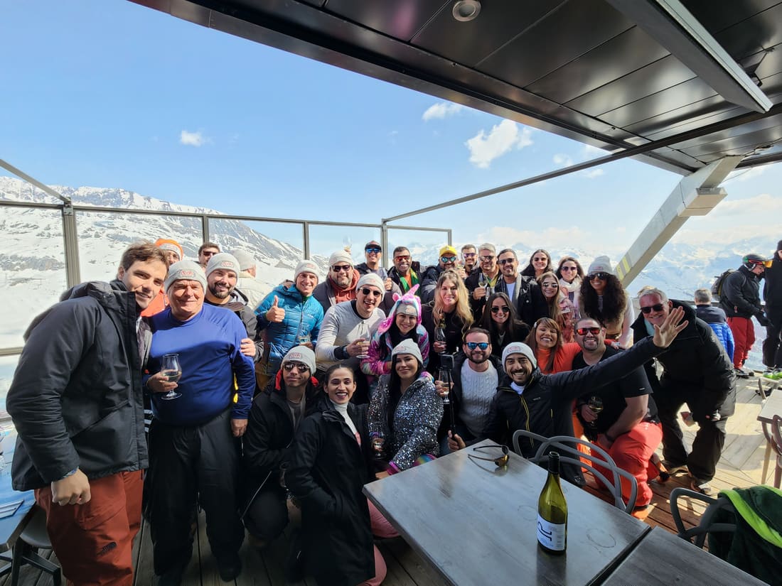 Grupo da Never Ends reunido no terraço de um restaurante de montanha em Alpe d'Huez, de toucas da agência, com os Alpes nevados pela vidraça