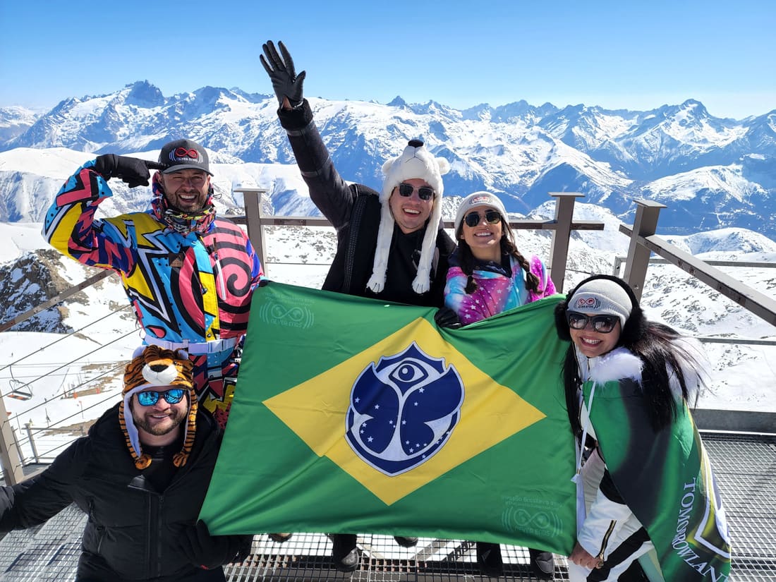 Viajantes da Never Ends no mirante do alto da montanha com a bandeira do Brasil estampada com o símbolo da Tomorrowland, e os picos dos Alpes franceses atrás