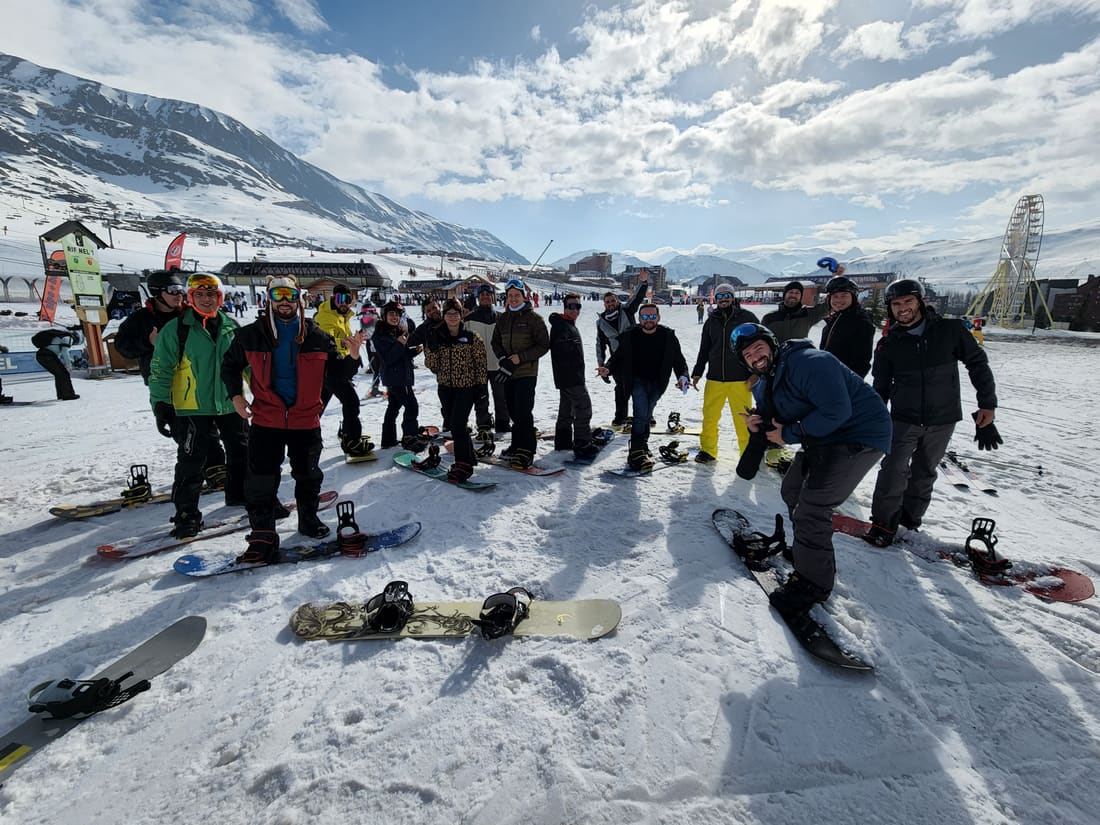 Grupo da Never Ends de pé com esquis e pranchas de snowboard na base da estação de Alpe d'Huez, com a roda gigante da vila ao fundo