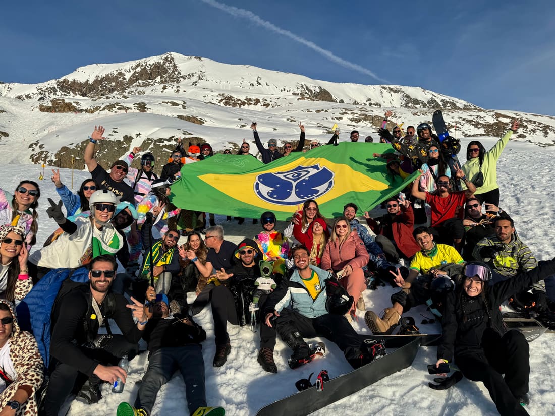 Grupo grande da Never Ends abrindo a bandeira gigante do Brasil com o símbolo da Tomorrowland na neve, com o pico de Alpe d'Huez atrás
