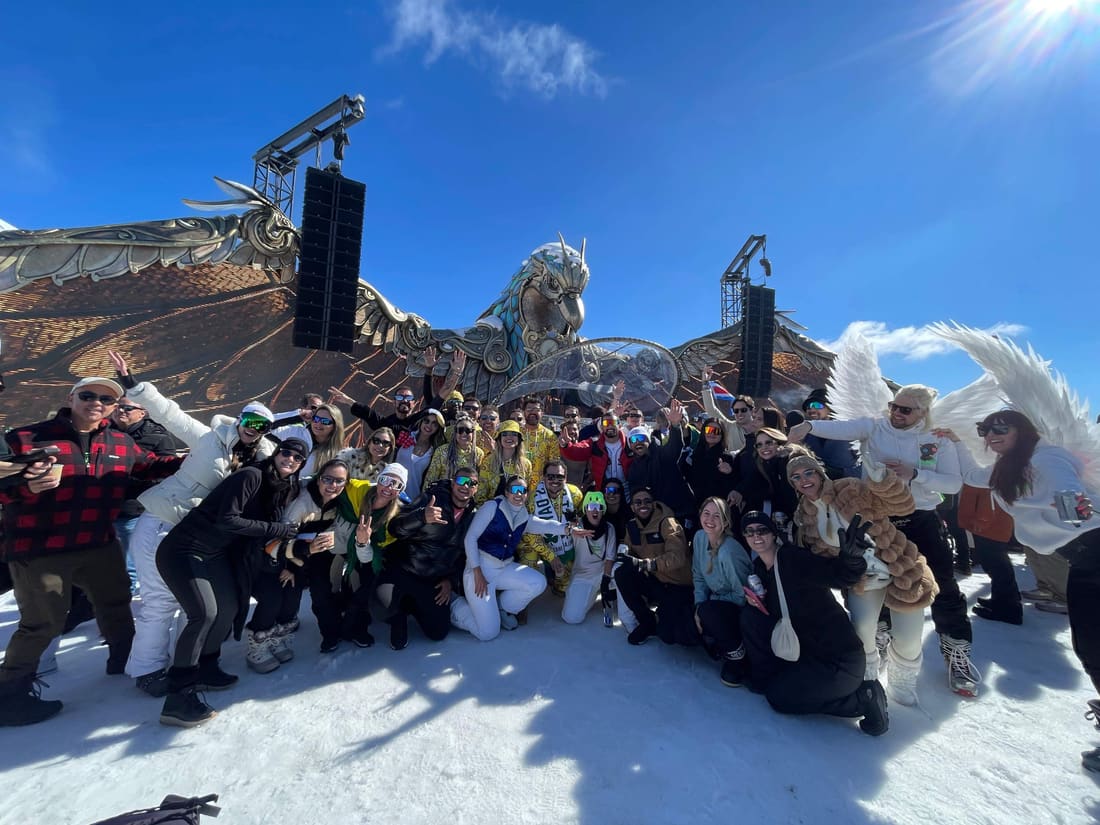 Grupo grande da Never Ends na neve diante do palco Paperworld da Tomorrowland Winter, com a ave esculpida em papel dourado, em Alpe d'Huez