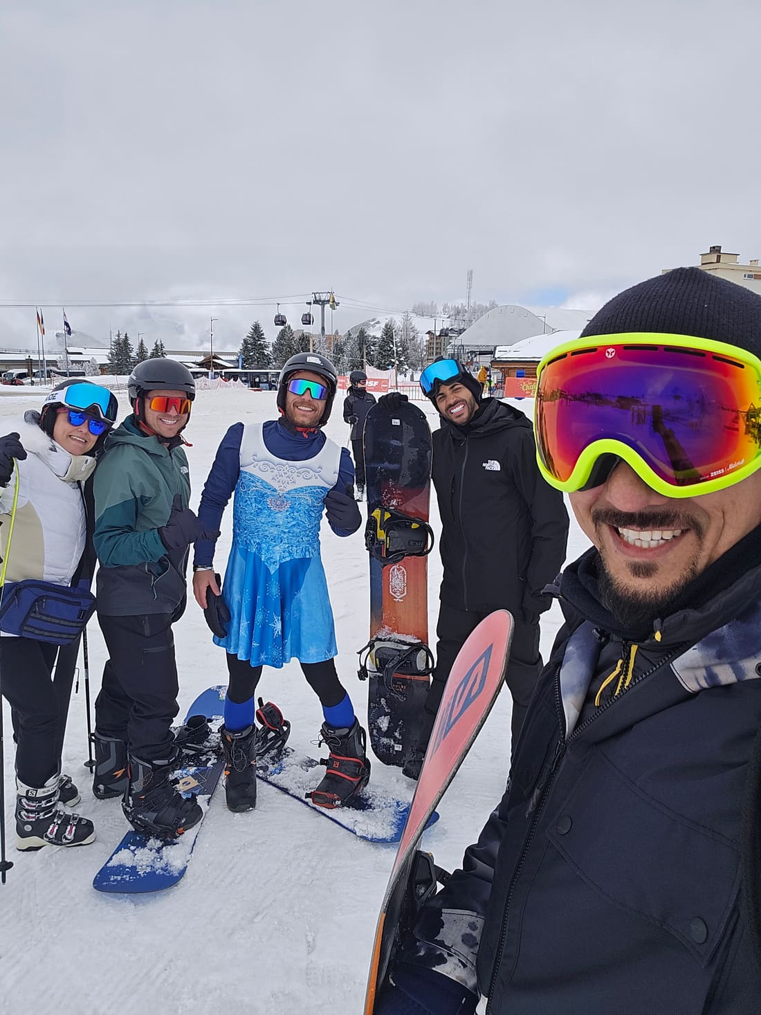 Viajantes da Never Ends com esquis e pranchas de snowboard na base da pista de Alpe d'Huez, com as gôndolas do teleférico ao fundo