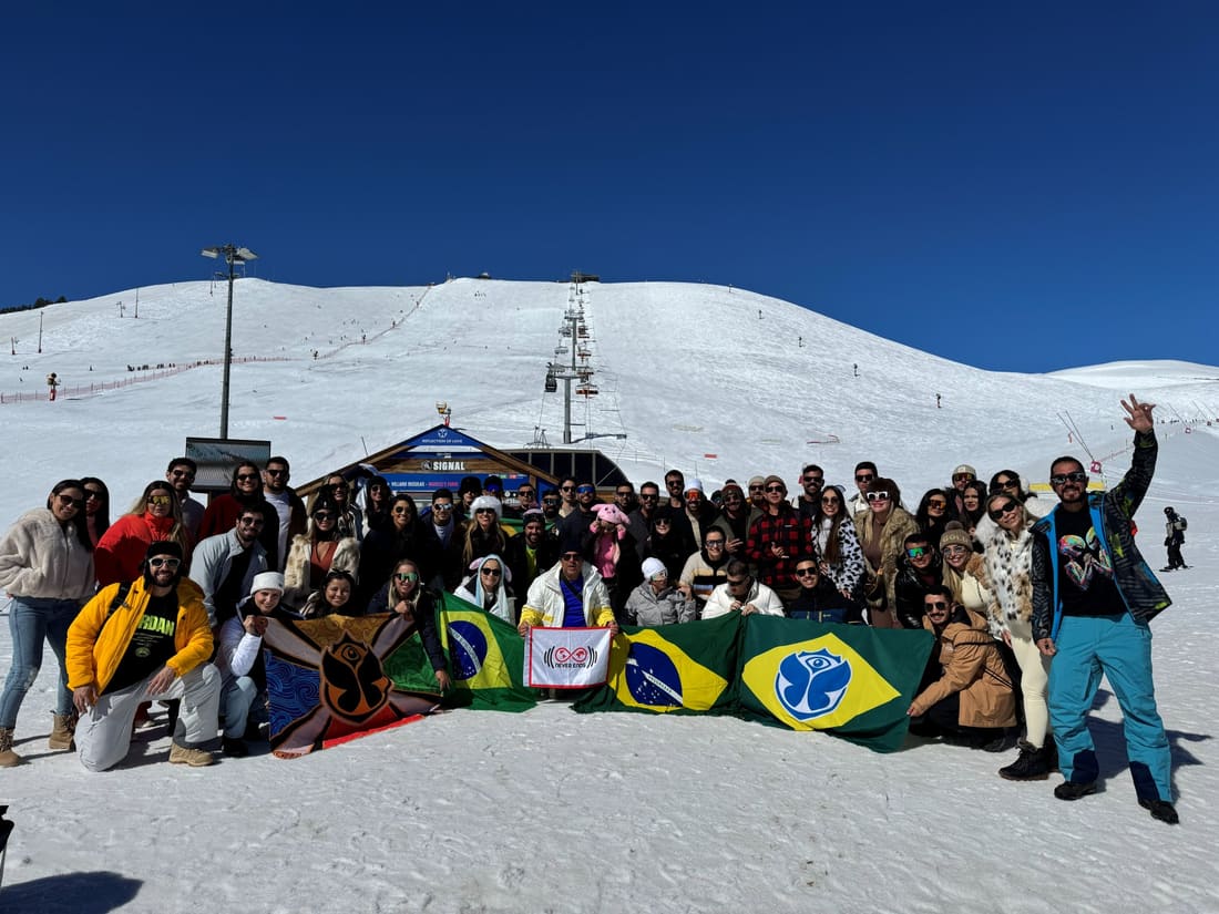 Grupo grande da Never Ends na neve diante do teleférico Signal, em Alpe d'Huez, com as bandeiras da agência, do Brasil e da Tomorrowland abertas