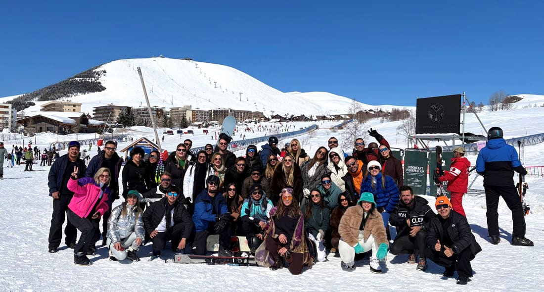 Grupo grande da Never Ends reunido na neve da base da estação de Alpe d'Huez, com a vila e as pistas de esqui atrás