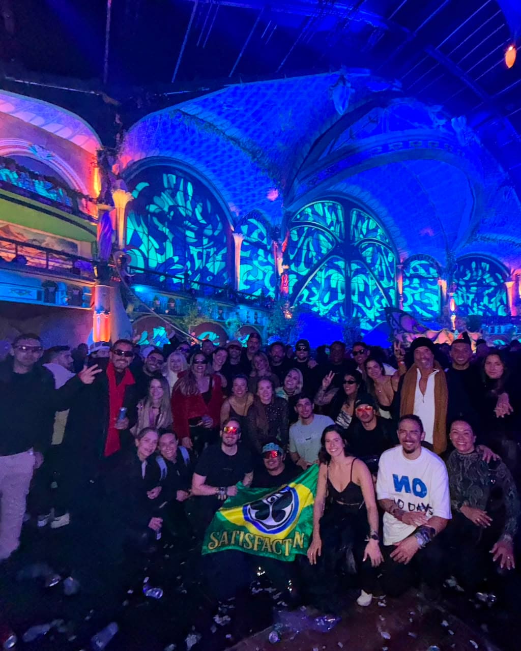Grupo da Never Ends com a bandeira do Brasil e o símbolo da Tomorrowland dentro do Mainstage coberto, iluminado de azul