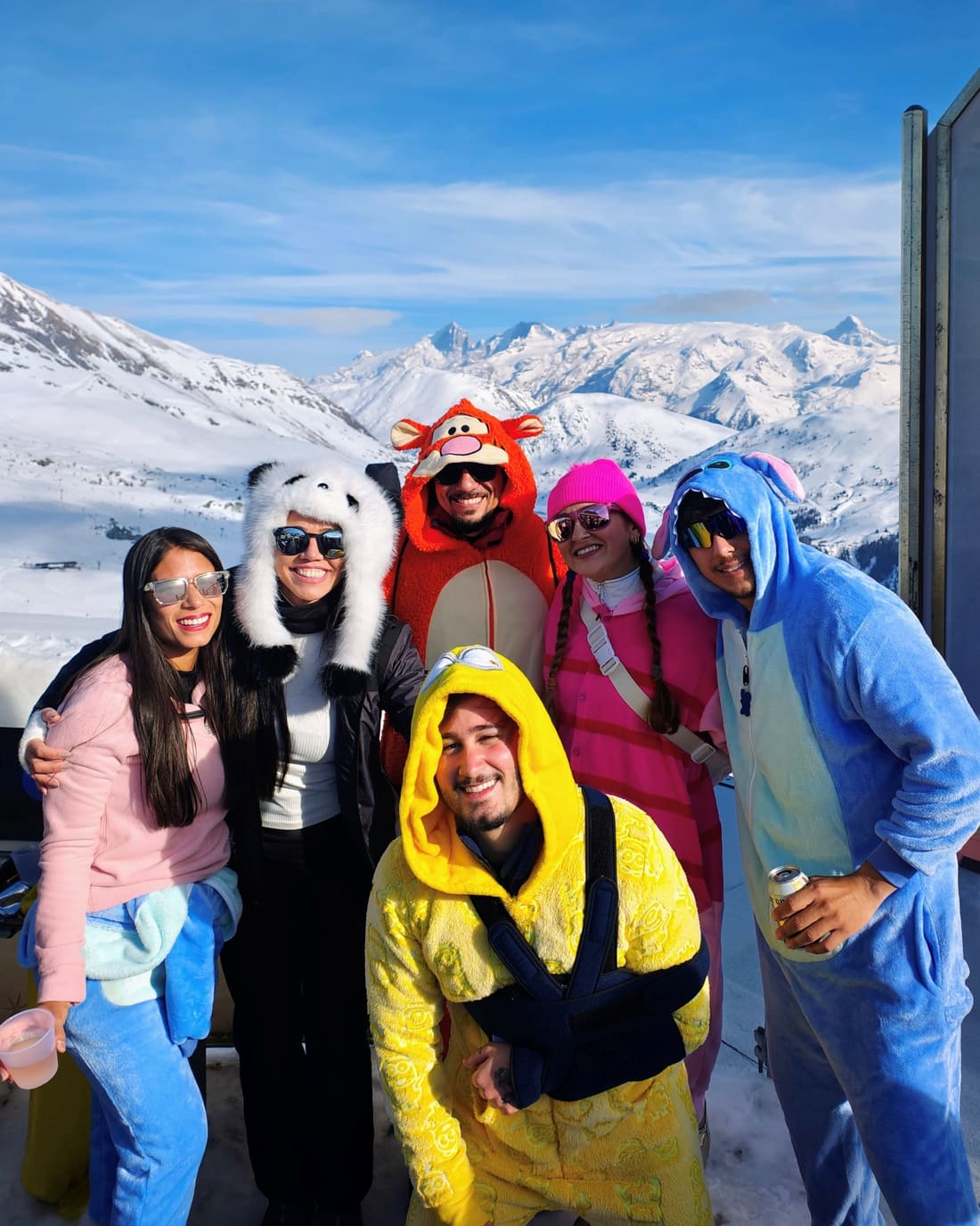 Viajantes da Never Ends de macacão colorido num terraço de Alpe d'Huez, com os Alpes franceses nevados ao fundo