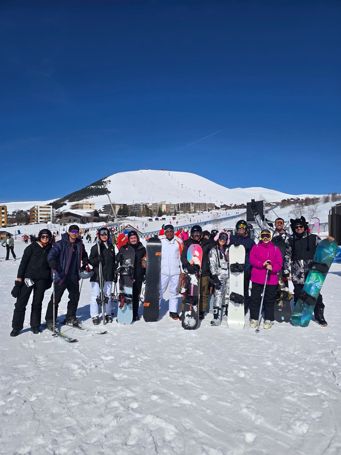Viajantes da Never Ends de pé com esquis e pranchas de snowboard na base da pista de Alpe d'Huez, com a montanha nevada atrás