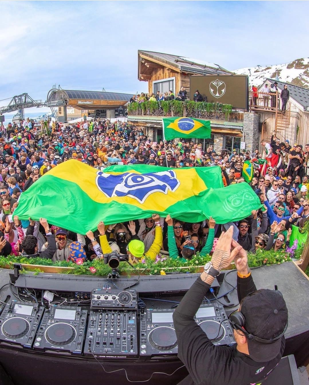 Público brasileiro erguendo a bandeira gigante do Brasil com o símbolo da Tomorrowland diante da cabine do DJ, num palco ao ar livre da Tomorrowland Winter em Alpe d'Huez