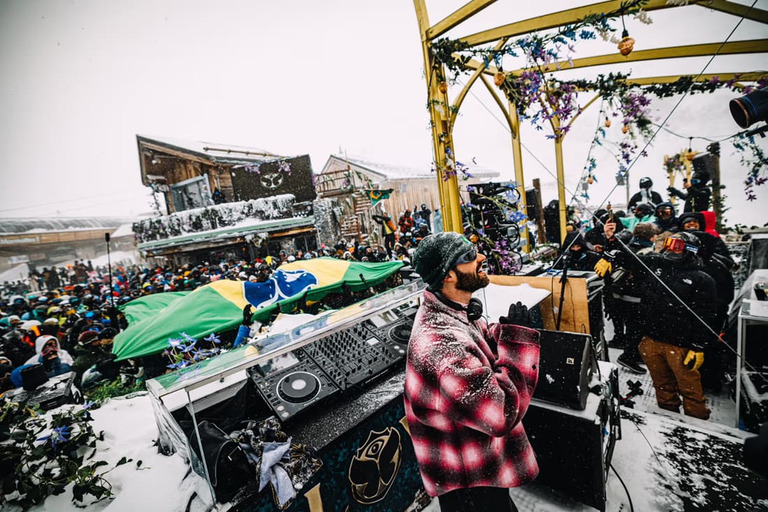DJ tocando debaixo de nevasca num palco da Tomorrowland Winter, com a bandeira do Brasil aberta pelo público brasileiro